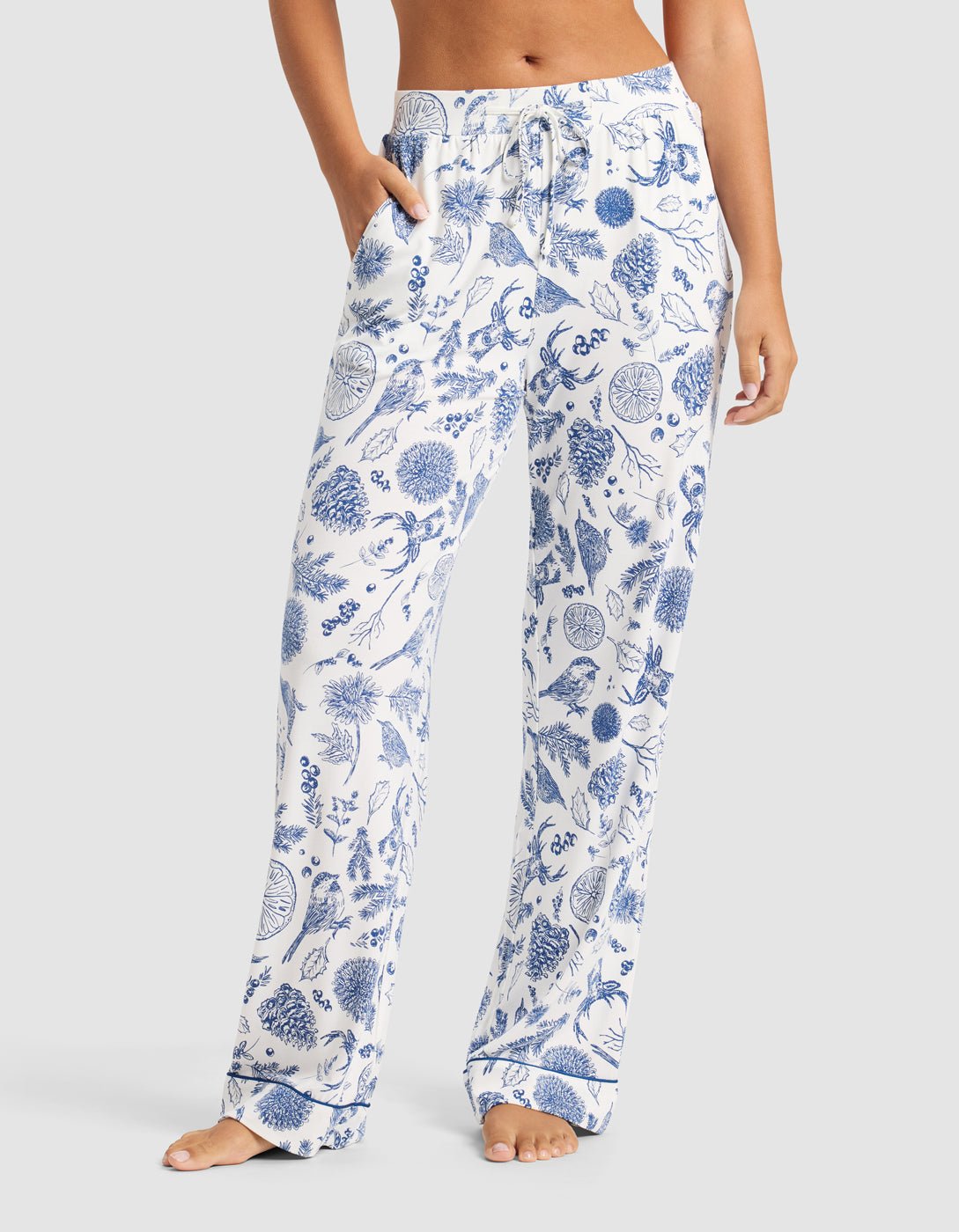 A woman wears Cozy Earth’s Women's Bamboo Stretch-Knit Pajama Pant in white with a blue botanical print, standing barefoot with hands in pockets against a plain background.|Color:Winter Toile Pacific