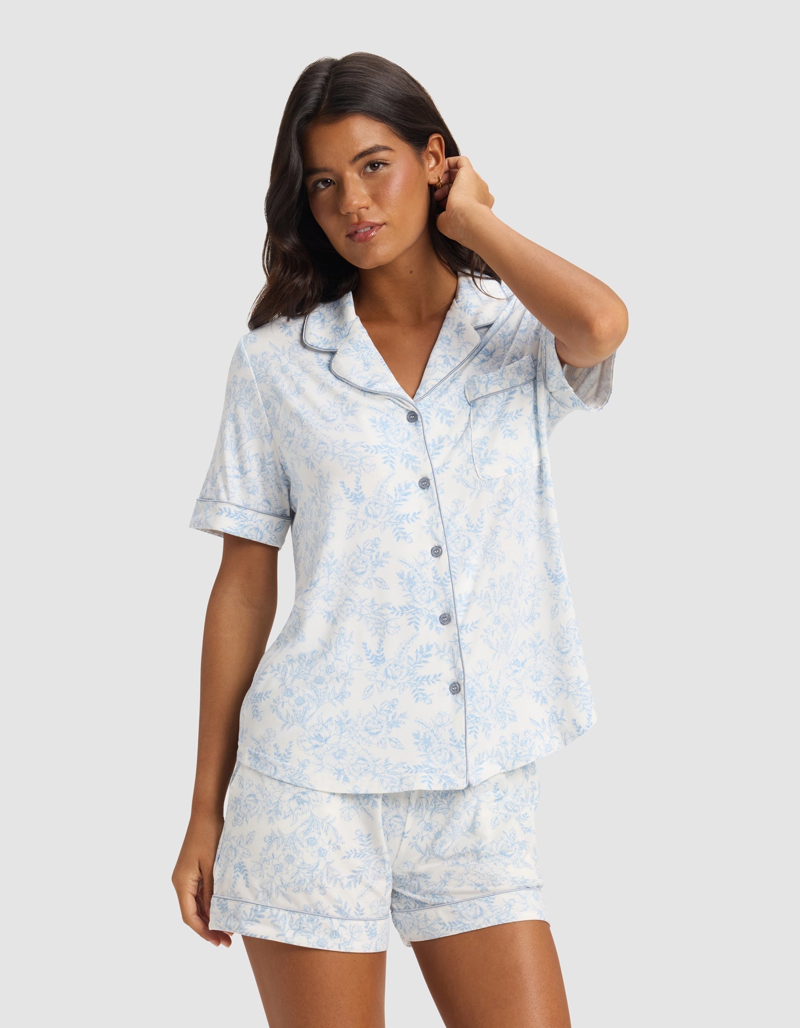 A woman with long dark hair models the Cozy Earth Women's Bamboo Stretch-Knit Short Sleeve Pajama Top in light blue and white with a floral pattern and buttons, standing against a plain light gray background.|Color:Sky Toile