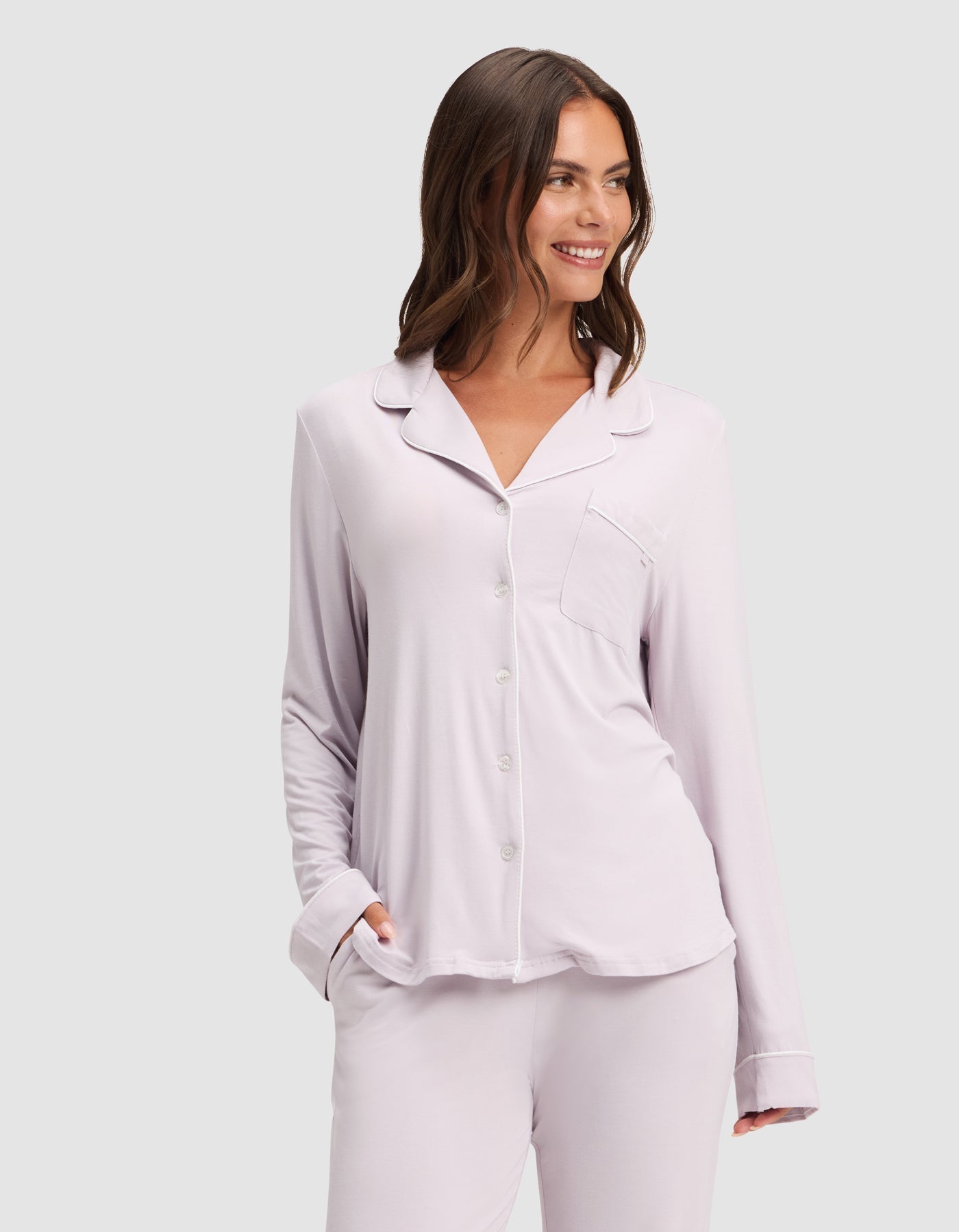 A woman with wavy brown hair smiles and looks to the side, wearing the HIDE Women's Bamboo Stretch-Knit Long Sleeve Pajama Top in light lavender, with her hand in her pocket, standing against a plain light background.  |Color:Lilac