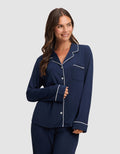 A woman with long brown hair smiles in a Cozy Earth Women's Bamboo Stretch-Knit Long Sleeve Pajama Top, navy blue with white piping, paired with matching pants, against a plain light gray background.  |Color:Navy