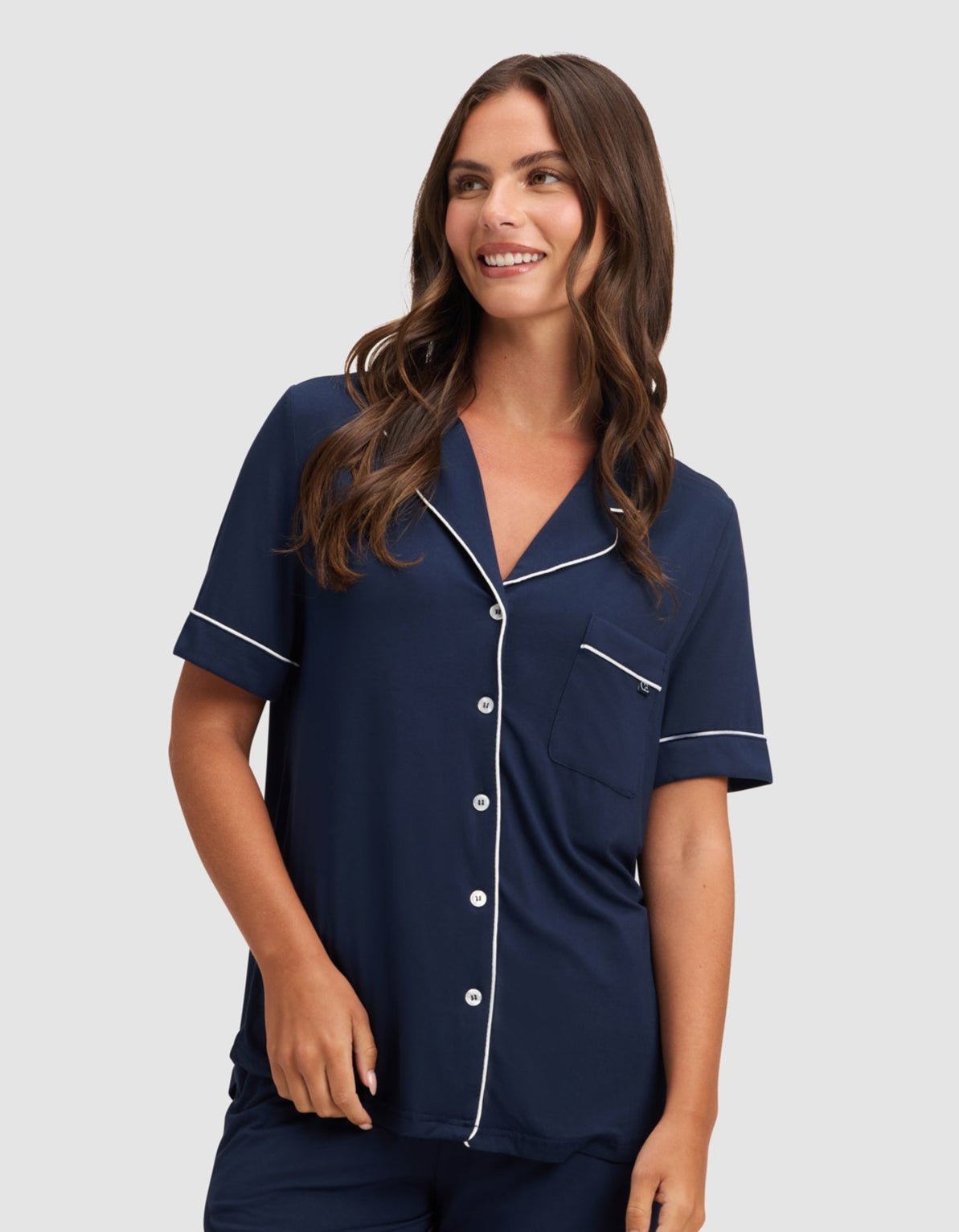 A woman smiles against a light gray background wearing HIDE's Women's Bamboo Stretch-Knit Short Sleeve Pajama Top in navy blue with white trim, paired with matching shorts. |Color:Navy